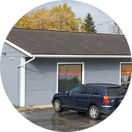 A dark blue SUV is parked in front of a one-story gray building with large windows, home to a local cannabis shop. Autumn trees with yellow leaves are visible in the background, and the pavement appears wet.