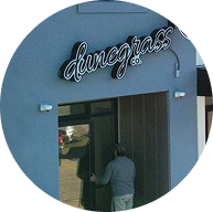 A person stands at the entrance of a gray building with a sign above the door that reads dunegrass in cursive letters, hinting at its connection to premium cannabis.