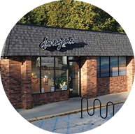 A small brick building with large front windows, a black shingled roof, and a sign reading “Boulevard” above the entrance. A bike rack and handicap parking spot sit out front, with trees and dunegrass visible in the background.
