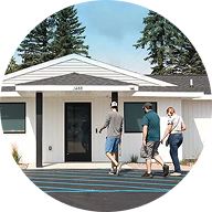 Three people are walking toward the entrance of a small, white, single-story building with a dark front door and large windows, surrounded by dunegrass, trees, and a clear sky.